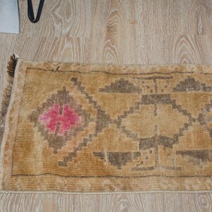 Puede incluir: Una alfombra rectangular de estilo vintage con fondo beige y patrones geom&eacute;tricos en marr&oacute;n y rosa. La alfombra tiene un borde con flecos y est&aacute; colocada sobre un suelo de madera clara.