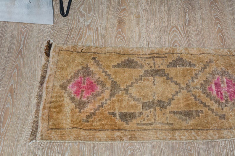 Puede incluir: Una alfombra rectangular de estilo vintage con fondo beige y patrones geom&eacute;tricos en marr&oacute;n y rosa. La alfombra tiene un borde con flecos y est&aacute; colocada sobre un suelo de madera clara.