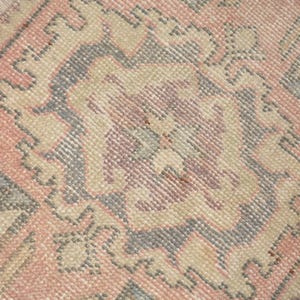 May include: A woven rug featuring a repeating geometric pattern in shades of muted pink, soft beige, and slate gray. The textile displays intricate symmetrical motifs with small floral-like accents in a darker pink hue throughout the design.