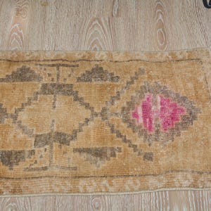 Puede incluir: Una alfombra rectangular de estilo vintage con fondo beige y patrones geom&eacute;tricos en marr&oacute;n oscuro y rosa. La alfombra tiene un aspecto desgastado, con desgaste visible, lo que aumenta su encanto antiguo. La alfombra est&aacute; colocada sobre un suelo de madera clara.