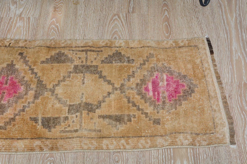 Puede incluir: Una alfombra rectangular de estilo vintage con fondo beige y patrones geom&eacute;tricos en marr&oacute;n oscuro y rosa. La alfombra tiene un aspecto desgastado, con desgaste visible, lo que aumenta su encanto antiguo. La alfombra est&aacute; colocada sobre un suelo de madera clara.