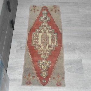 May include: A rectangular, vintage-style rug with a central diamond pattern in red and beige. The rug has a grey border and floral accents at the top and bottom. The rug is placed on a light grey wooden floor.