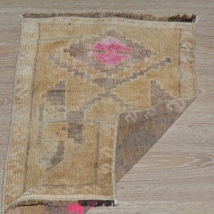Puede incluir: Una peque&ntilde;a alfombra rectangular con un patr&oacute;n geom&eacute;trico. La alfombra presenta una forma de diamante central en rosa, rodeada de tonos marrones y beige. Los bordes est&aacute;n deshilachados y la alfombra est&aacute; colocada sobre una superficie de madera.