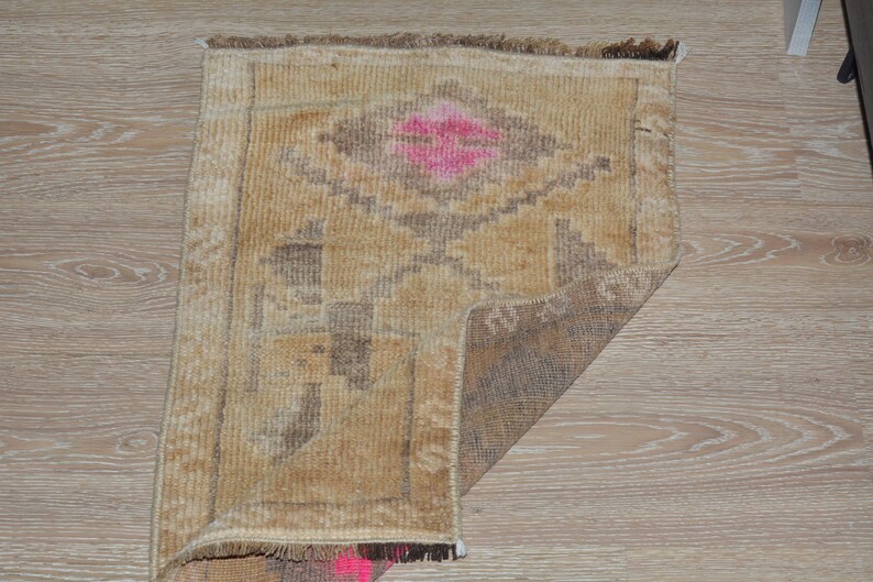 Puede incluir: Una peque&ntilde;a alfombra rectangular con un patr&oacute;n geom&eacute;trico. La alfombra presenta una forma de diamante central en rosa, rodeada de tonos marrones y beige. Los bordes est&aacute;n deshilachados y la alfombra est&aacute; colocada sobre una superficie de madera.