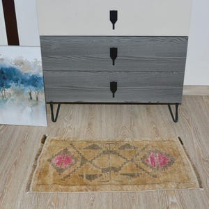 Puede incluir: Una peque&ntilde;a alfombra rectangular con un patr&oacute;n geom&eacute;trico y detalles florales rosas. La alfombra es de color beige con un dise&ntilde;o marr&oacute;n y gris. Se coloca sobre un suelo de madera clara frente a una c&oacute;moda de tres cajones.
