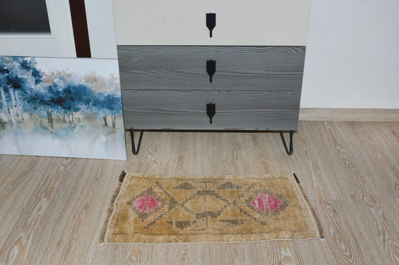 Puede incluir: Una peque&ntilde;a alfombra rectangular con un patr&oacute;n geom&eacute;trico y detalles florales rosas. La alfombra es de color beige con un dise&ntilde;o marr&oacute;n y gris. Se coloca sobre un suelo de madera clara frente a una c&oacute;moda de tres cajones.