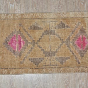 Puede incluir: Alfombra rectangular beige con un patr&oacute;n geom&eacute;trico. La alfombra presenta un dise&ntilde;o central de diamantes y tri&aacute;ngulos en tonos marr&oacute;n y rosa. Tiene un borde con flecos y est&aacute; sobre una superficie de madera clara.