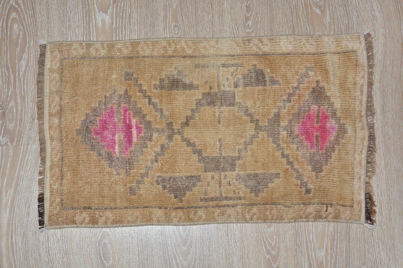 Puede incluir: Alfombra rectangular beige con un patr&oacute;n geom&eacute;trico. La alfombra presenta un dise&ntilde;o central de diamantes y tri&aacute;ngulos en tonos marr&oacute;n y rosa. Tiene un borde con flecos y est&aacute; sobre una superficie de madera clara.