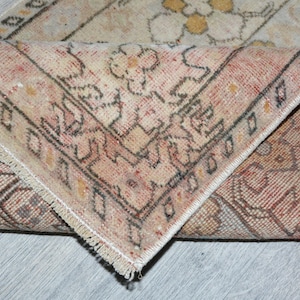 Pode incluir: Secção de um tapete vintage com um canto dobrado, revelando a parte inferior. O tapete apresenta um design desbotado e desgastado em tons de bege, vermelho e castanho, com padrões florais e geométricos. As bordas são finalizadas com franjas.