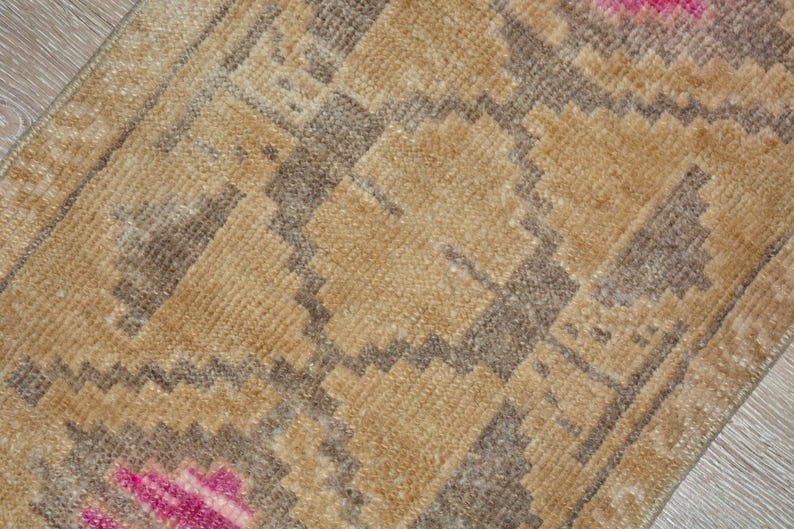 Puede incluir: Primer plano de una peque&ntilde;a alfombra rectangular con un patr&oacute;n geom&eacute;trico. La alfombra presenta un dise&ntilde;o repetitivo en tonos beige, marr&oacute;n y rosa. La textura parece suave y tejida, lo que sugiere un estilo hecho a mano o tradicional.