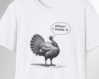May include: White t-shirt featuring a detailed illustration of a turkey with its feathers fanned out. A speech bubble above the turkey reads "phew! I made it". The design is in black and white.