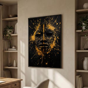 May include: Black framed art print featuring a stylized mask with a textured surface. The mask is black with gold paint splatter details. The artwork is displayed on a neutral-colored wall.
