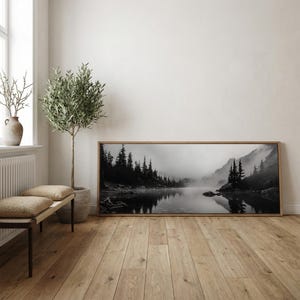 May include: A black and white landscape photograph of a misty lake surrounded by evergreen trees and mountains. The artwork is displayed in a rectangular wooden frame, leaning against a white wall. The room features a wooden bench, a potted tree, and a vase.