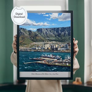Puede incluir: Una impresión enmarcada de la Montaña de la Mesa en Ciudad del Cabo, Sudáfrica, con un paisaje urbano y puerto vibrante bajo un cielo azul con nubes. La obra de arte es una descarga digital.