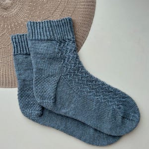 Könnte beinhalten: Ein Paar blaue Stricksocken mit einem strukturierten Rautenmuster auf dem Fußrücken. Die Socken haben einen gerippten Bund und sind aus einem weichen, möglicherweise wollenen Material gefertigt. Die Socken liegen auf einem beigefarbenen, gewebten Platzset.