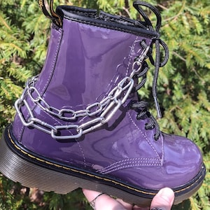 May include: A shiny, purple combat boot with a silver chain detail. The boot has black laces and a black sole. The image is taken outdoors with green foliage in the background.