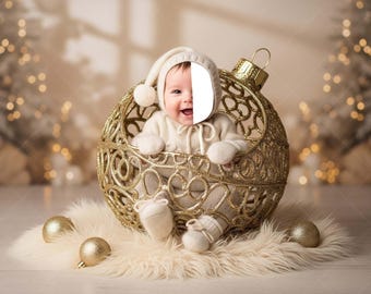 May include: A baby dressed in a cream-colored outfit and hat sits inside a large, ornate gold Christmas ornament. The ornament rests on a fluffy, cream-colored rug with gold ornaments scattered around it. The background is softly blurred with Christmas lights.
