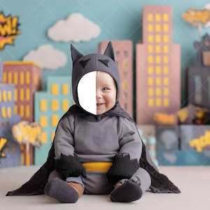 May include: A baby dressed in a gray Batman costume, including a cowl, cape, and gloves, sits in front of a cityscape backdrop. The costume has a yellow belt. Comic book-style "POW!" graphics are in the background.
