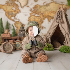 Puede incluir: Un bebé con un mono caqui y un sombrero de camuflaje, sosteniendo unos prismáticos. El fondo presenta un mapa del mundo, un pequeño tipi y un globo terráqueo. La escena evoca un tema de viaje o aventura. El bebé lleva guantes verdes y botas marrones.