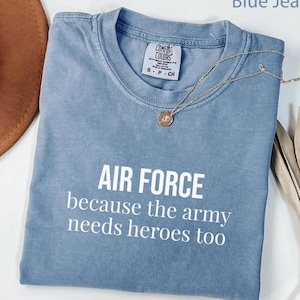 Op de afbeelding: Een blauw jeanskleurig t-shirt met witte tekst die luidt AIR FORCE because the army needs heroes too. Het shirt heeft een ronde hals en een Comfort Colors merklabel aan de binnenkant van de kraag. Een dunne gouden ketting ligt op de stof.