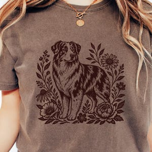 May include: An espresso-colored t-shirt featuring a dark brown graphic print of an Australian Shepherd dog surrounded by various flowers and leaves. The shirt has a crew neckline and short sleeves, with the word Espresso printed in the top corner.