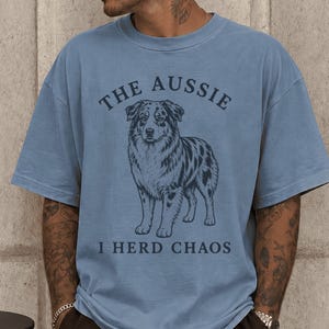 May include: A blue jean coloured short-sleeve t-shirt featuring a dark blue graphic of an Australian Shepherd dog in the centre. The text above the dog reads THE AUSSIE and the text below the dog reads I HERD CHAOS in a dark blue font.