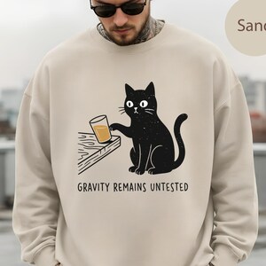 Puede incluir: Una sudadera de cuello redondo color arena con un gráfico de un gato negro sentado en el borde de una mesa, empujando un vaso de líquido naranja. Debajo del gato, el texto dice: "GRAVITY REMAINS UNTESTED". Tiene mangas largas y cuello acanalado.