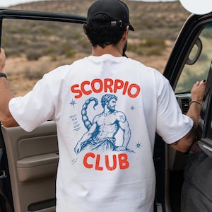 以下が含まれることがあります： 背面に赤い文字でSCORPIO CLUBとプリントされた白いTシャツ。デザインには、サソリの尾を持つ筋肉質な人物の青いイラスト、TRUST FEW CUT DEEPという文字、小さな星のアクセントが含まれています。