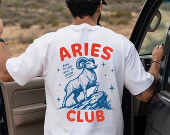 May include: A white t-shirt featuring a graphic on the back with the red text ARIES CLUB and a blue illustration of a ram standing on a rock. The text BORN TO LEAD. BUILT TO ENDURE. is printed in blue next to the ram, surrounded by small blue stars.