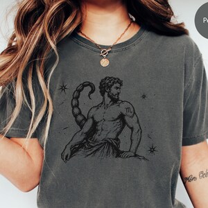 Puede incluir: Una camiseta gris carbón con una ilustración negra estilo tinta de una figura musculosa con cola de escorpión y el símbolo del zodiaco Escorpio en el hombro. El diseño incluye pequeños detalles de estrellas y el texto Pepper en la esquina.