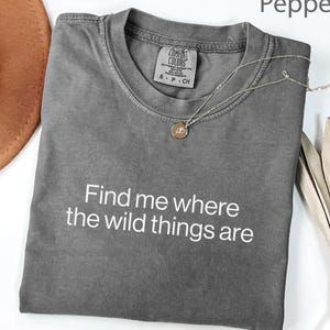 May include: A charcoal gray short-sleeve t-shirt with a crew neckline and the white printed text "Find me where the wild things are" across the chest. The shirt features a Comfort Colors brand label at the inner neck and a thin gold necklace.
