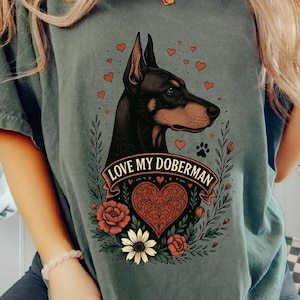 May include: A slate gray short-sleeve t-shirt featuring a graphic of a Doberman dog profile surrounded by small red hearts, green leaves, and flowers. Below the dog, a banner contains the text LOVE MY DOBERMAN above a large decorative heart.