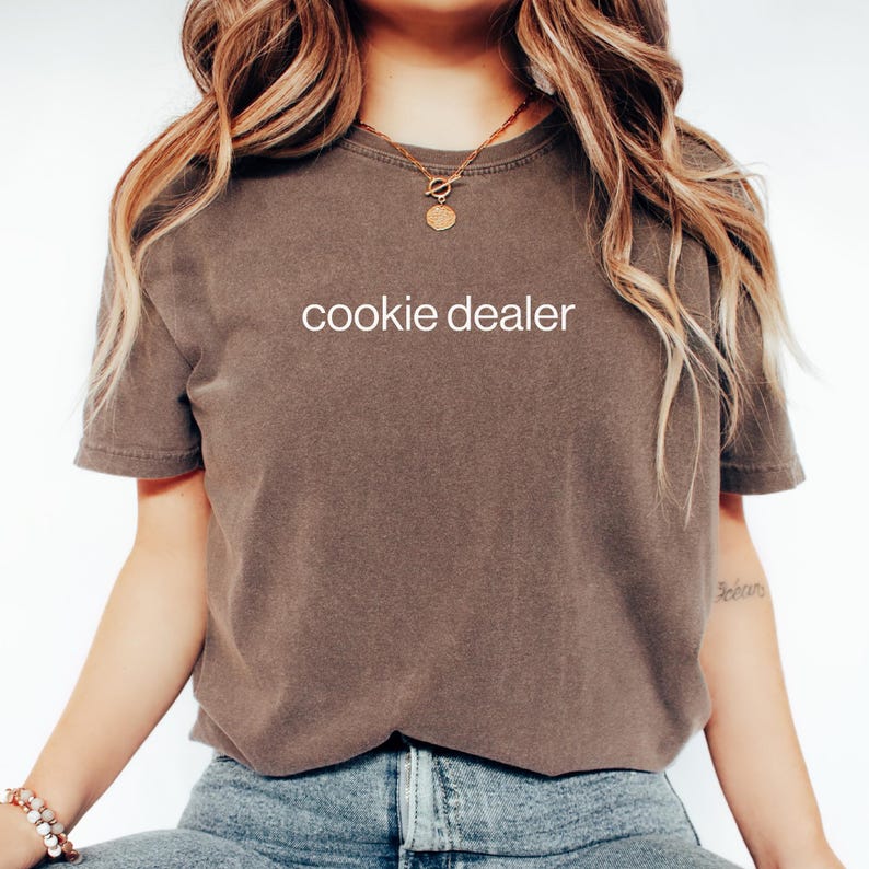 May include: A brown t-shirt with the words "cookie dealer" printed in white. The shirt is a solid color and has short sleeves. The model is wearing blue jeans and a gold necklace.