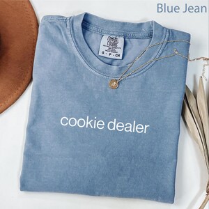May include: A blue jean Comfort Colors t-shirt with the text "cookie dealer" in white. A gold necklace with a pendant is draped across the shirt. A brown hat and dried plant stems are in the background.