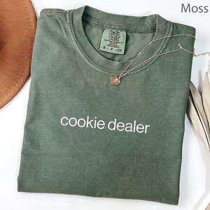 May include: A moss green t-shirt with the words "cookie dealer" printed in white. The shirt has a crew neck and short sleeves. A gold-colored necklace with a round pendant is draped on the shirt. The shirt is labeled "Comfort Colors".
