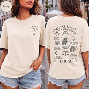 Könnte beinhalten: Ein elfenbeinfarbenes Kurzarm-T-Shirt mit einer kleinen Waage-Sternzeichen-Grafik auf der linken Brust. Die Rückseite zeigt eine große Grafik mit dem Text Balance, Beauty & Just A Bit Of Drama, Libra und verschiedenen Symbolen wie Waagen, Mond und Blumen.