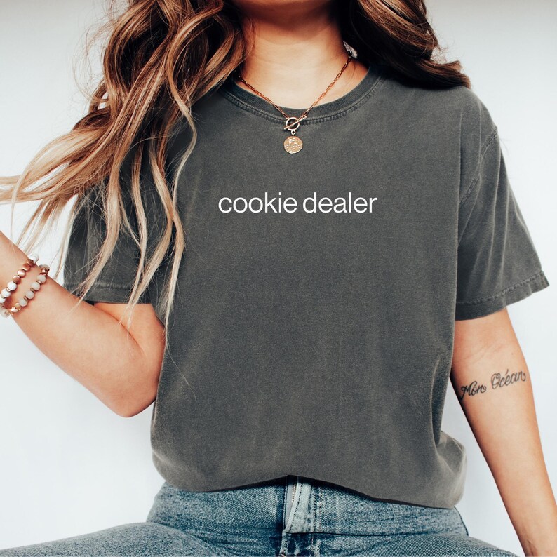 May include: Dark gray t-shirt with the text "cookie dealer" in white. The shirt is a relaxed fit and has a crew neck. The model is wearing blue jeans and a gold necklace.