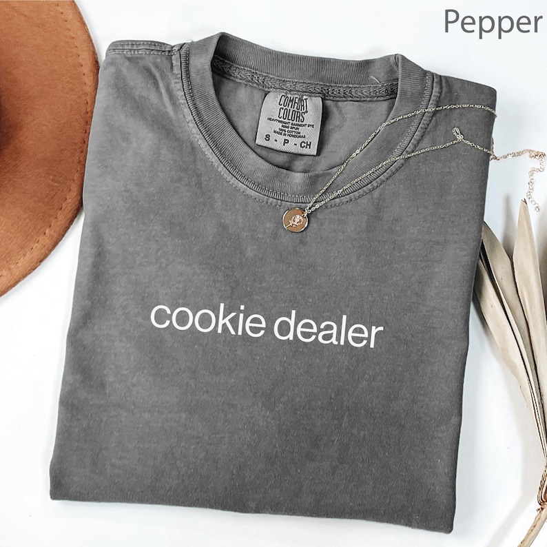 May include: A gray Comfort Colors t-shirt with the words "cookie dealer" printed in white. A silver necklace with a small pendant is draped across the shirt. The shirt is folded on a white surface.