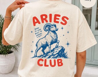 May include: An ivory t-shirt featuring a back graphic with the red text ARIES CLUB and a blue illustration of a ram standing on a rock. The design includes blue stars and the text BORN TO LEAD. BUILT TO ENDURE. The shirt has short sleeves.