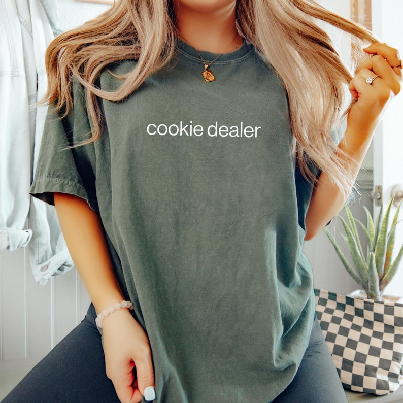 May include: A dark green t-shirt with the text "cookie dealer" in white. The shirt is a relaxed fit and appears to be made of a soft material. The model is wearing the shirt with dark pants.