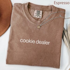 May include: Espresso-colored t-shirt with the text "cookie dealer" in white. The shirt is folded and has a Comfort Colors tag. A silver necklace with a round pendant is draped across the shirt.