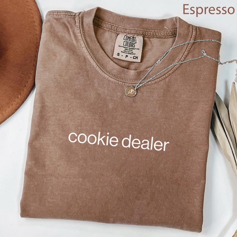 May include: Espresso-colored t-shirt with the text "cookie dealer" in white. The shirt is folded and has a Comfort Colors tag. A silver necklace with a round pendant is draped across the shirt.