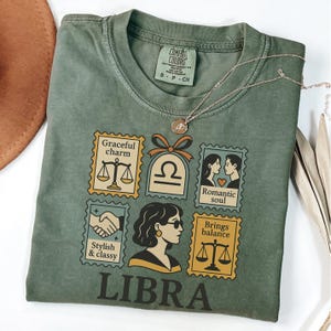 May include: Green Comfort Colors t-shirt featuring a Libra zodiac design. The design includes Libra symbols, illustrations, and the word "LIBRA". A silver necklace with a pendant is draped on the shirt.