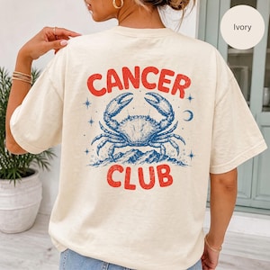 May include: An ivory t-shirt featuring a graphic on the back with the text "CANCER CLUB" in red letters. The design includes a blue illustration of a crab centred between the words, surrounded by small blue stars and a crescent moon symbol.