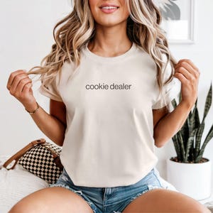 May include: Cream-colored t-shirt with the text "cookie dealer" in black. The shirt is a crew neck style and is being worn by a person. The person is wearing blue jean shorts. The background includes a plant and a decorative pillow.