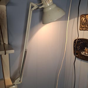 May include: A vintage, adjustable desk lamp in a light grey colour. The lamp has a large, conical shade and an articulated arm for flexible positioning. The lamp is plugged into a wall socket.
