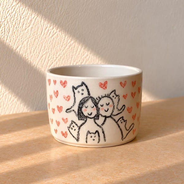 May include: A white ceramic mug featuring a hand-drawn black ink illustration of two people surrounded by several small cats. The design is accented with numerous small red hearts scattered across the exterior surface of the cylindrical vessel.