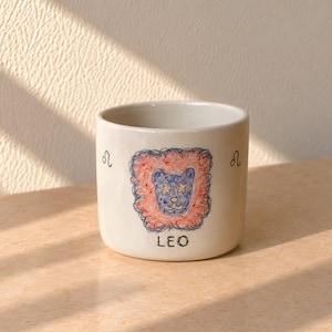 Puede incluir: Una taza de cerámica blanca con una ilustración dibujada a mano de una cara de león azul con ojos en forma de estrella, rodeada por una melena garabateada en rojo y azul. La palabra LEO está impresa debajo del león, flanqueada por dos símbolos.
