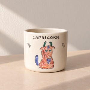 Puede incluir: Una taza de cerámica blanca con una ilustración dibujada a mano de una cabra de Capricornio en naranja, azul y verde. La palabra CAPRICORN está escrita en tinta negra sobre la cabra, con el símbolo del zodiaco Capricornio a ambos lados.