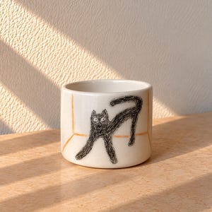 Op de afbeelding: Een witte keramische beker met een handgetekende zwarte schets van een kat met grote ogen die zich uitrekt. De illustratie is omlijst door dunne, oranje handgeschilderde lijnen die een hoekeffect creëren op het buitenoppervlak van de beker.